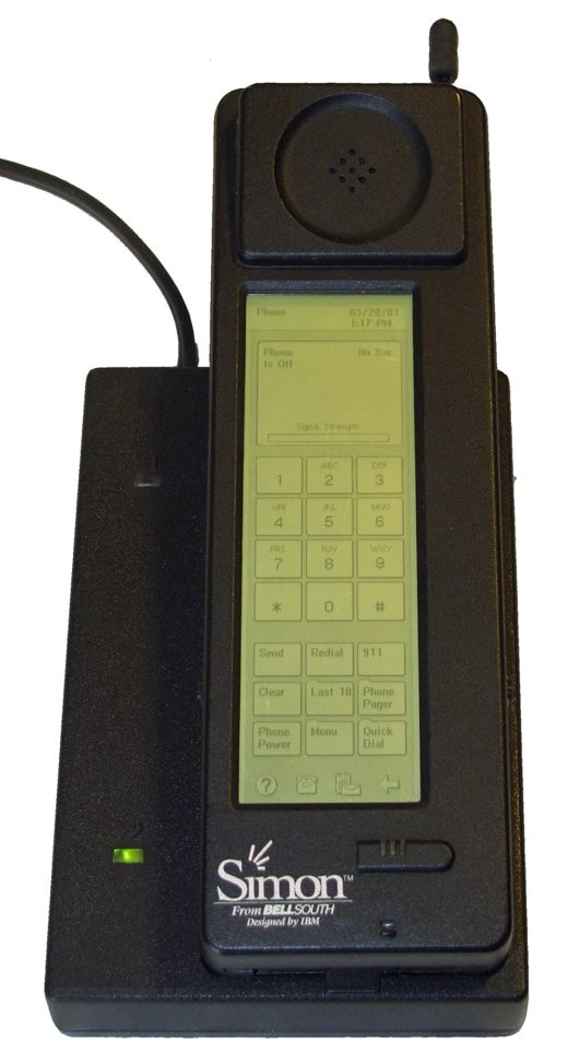A tall black IBM Simon handset with a monochrome touchscreen showing a phone keypad, standing in its charging base.