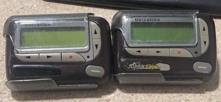 Two black alphanumeric pagers with small green LCD screens lying side by side on a counter.